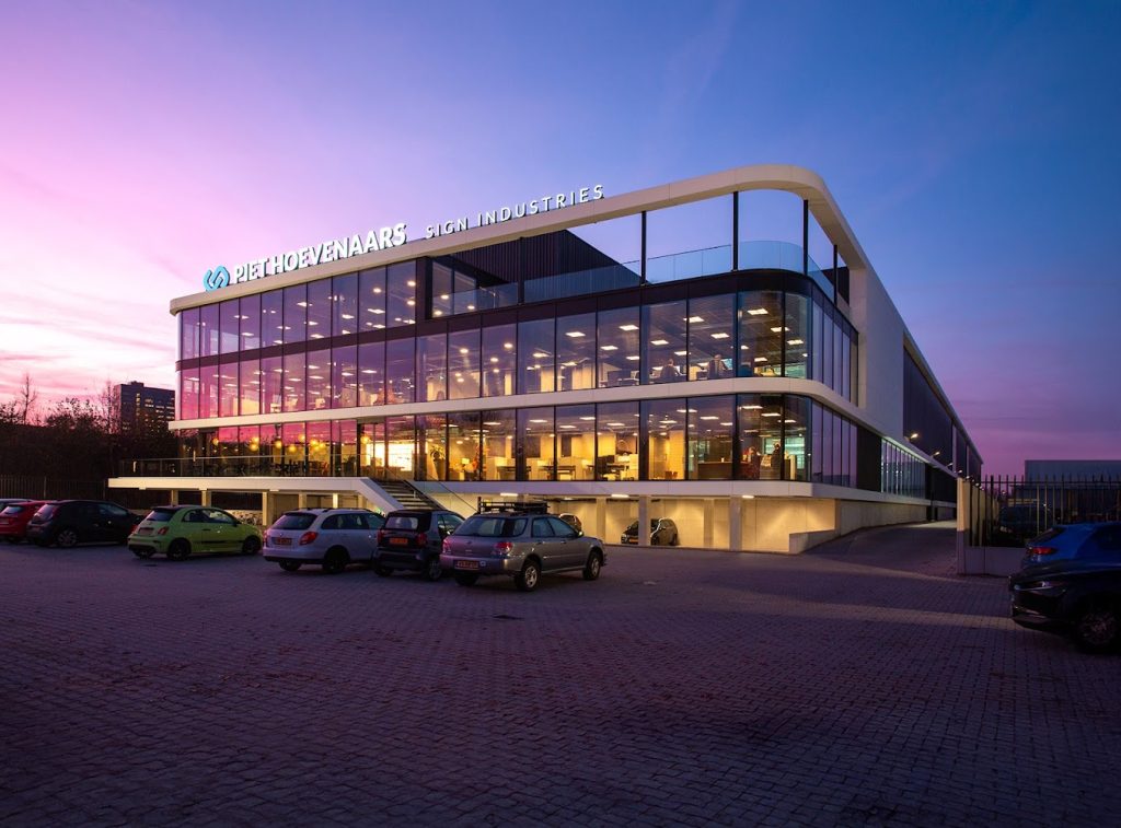 Glazen kantoorgebouw bij zonsondergang met parkeerplaats.