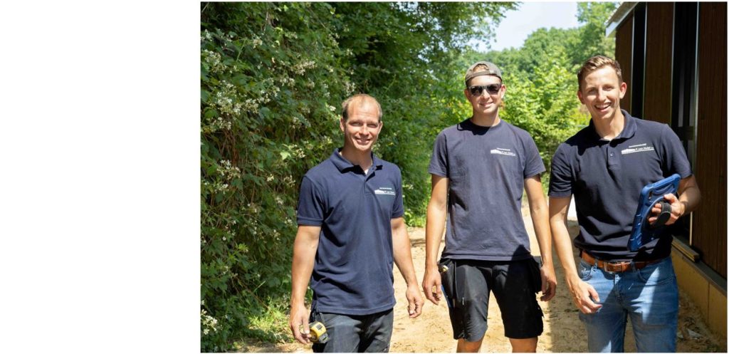Drie mannen in bedrijfskleding buiten bij groen struikgewas.